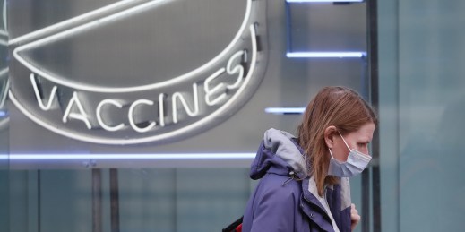 A woman wearing a mask walks past a neon sign reading "Vaccines."